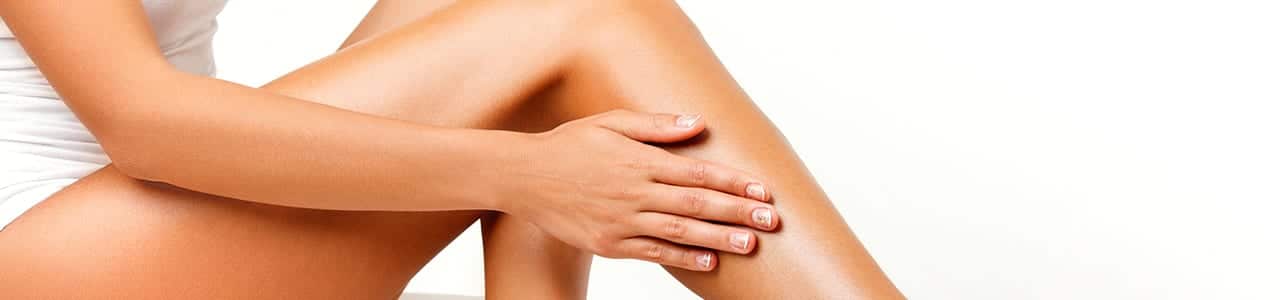 close up of woman's bare legs as she glides hand across calf in white t shirt with solid white background